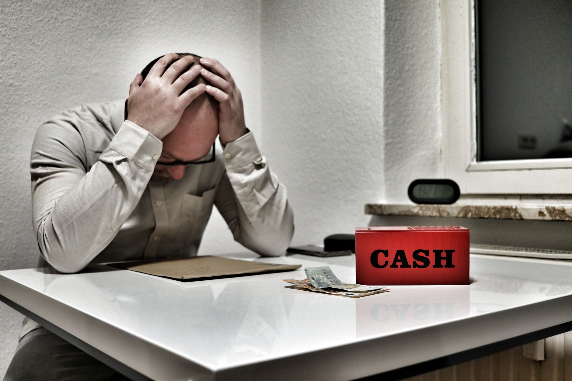 Person holding head in despair at desk with cashbox and money, representing financial stress.