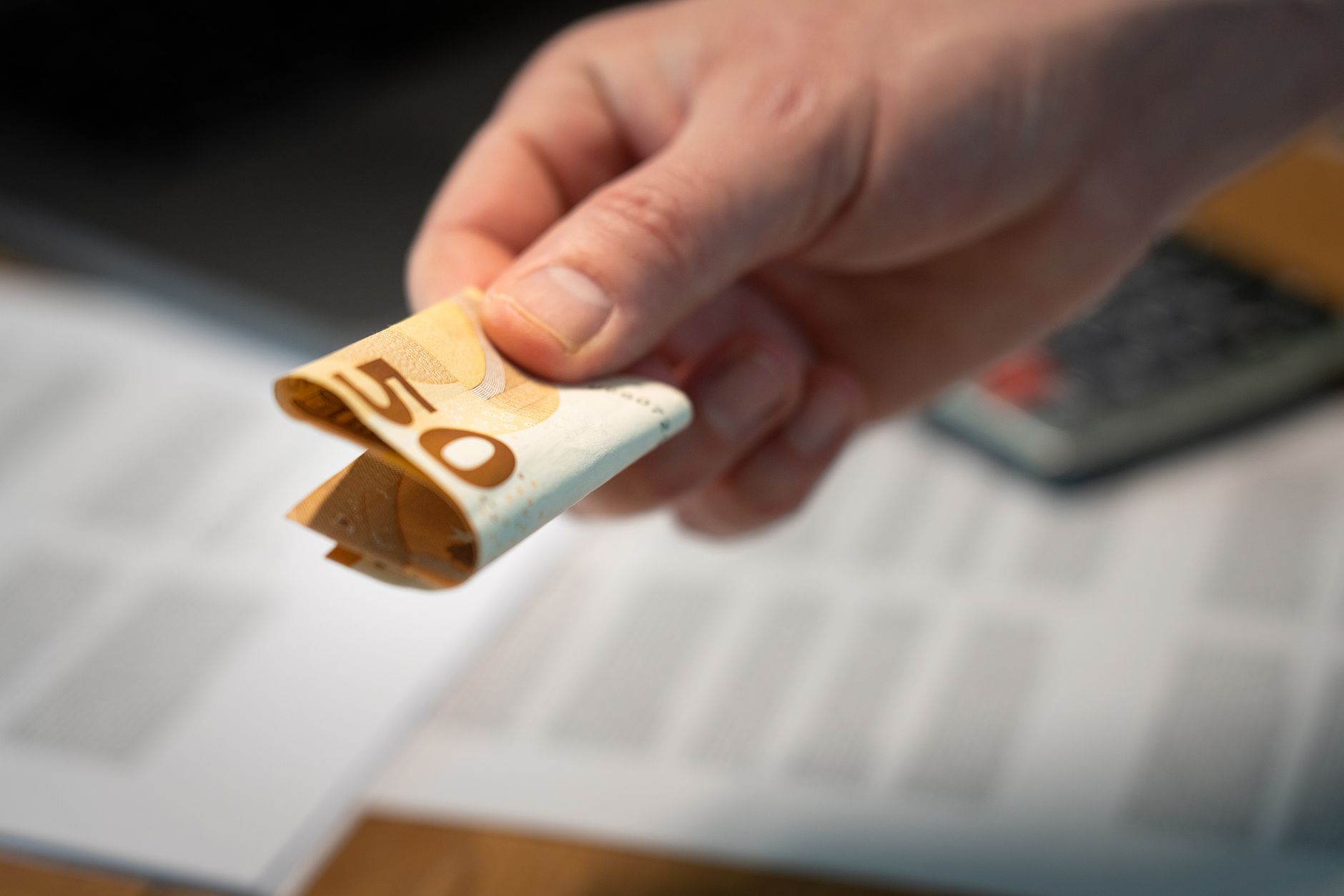 A hand holding a folded 50 euro banknote with documents in the background.